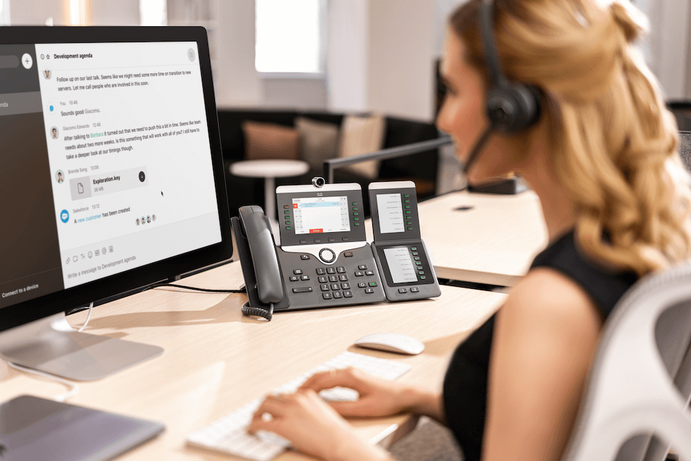 Eine Frau telefoniert über das Cisco IP-Telefon mit Videofunktion und ihr Headset, während sie Daten in den Computer einträgt